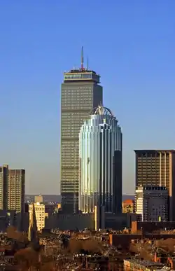 Distant aerial view of two skyscrapers in early evening; the tallest is a large, gray rectangular tower with a steel latticework facade and a tall antenna mast on its roof. The shorter skyscraper, directly in front of the other building, has a tinted, all-glass facade with a prominent, open-air dome. Several high-rises surround the two skyscrapers on all sides.