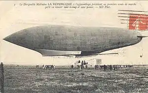 Postcard with orange stamp (French) affixed shows left flank of an airship just above the ground; some people are on the ground below the airship