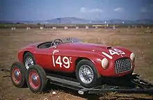 A sporty red two-seat convertible facing right at a mild three-quarter orientation to the viewer, parked in front of a light brown wall