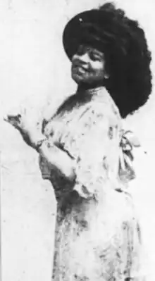 An African-American woman, standing and smiling with hands at her chest; she is wearing a light-colored gown and a large dark hat