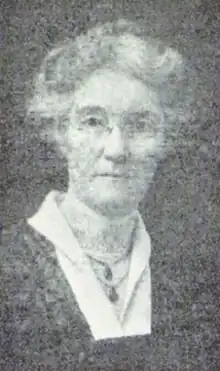 An older white woman with grey hair in a bouffant updo, wearing glasses and two pendants.