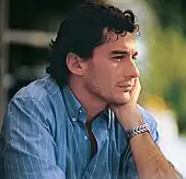 Photographic portrait of Ayrton Senna predominately showing the right side of his face with his left hand on the left hand side of his face and he is wearing a T-shirt