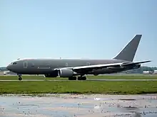 Gray jet aircraft facing left on apron against a cloudless, pale blue sky. In the foreground are green grass; the foreground is a wet tarmac.