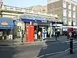 A single storey pale brick building topped with stone railing. Above an entrance is a canopy, around which are rectanglar blue signs reading "BAYSWATER" and "BAYSWATER STATION". People are walking in the street.