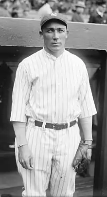 Three-quarter-length picture of a man in a stadium. He wears a striped baseball uniform, a cap with the NY logo, and a baseball glove. He has a long face and tanned complexion with deepset eyes under a heavy brow, and has a small full mouth. He looks out towards the field with a calculating expression, his eyes narrowed and his jaw clenched.