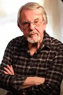 Picture of a white man with short grey hair, a short goatee, and cross arms wearing a flannel shirt