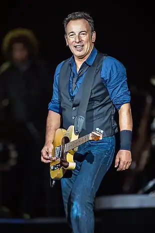 A man holding a guitar, wearing a blue shirt and a dark vest