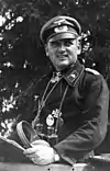 A man wearing a peaked cap, black military uniform with various military decorations.