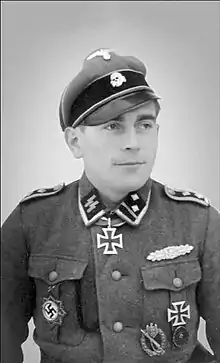 A man wearing a peaked cap with skull emblem, a military uniform with various military decorations and an Iron Cross displayed at the front of his uniform collar.