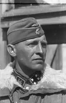 A black-and-white photograph of a man wearing a military uniform with fur collar, side cap and a neck order in shape of an Iron Cross. His cap has an emblem in shape of a human skull and crossed bones.