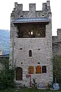 Tower in the centre of a stone wall. At the bottom left of the tower there is a door and then, on its right, two small wooden shutters. In the middle of the tower wall there is a small window and at the top of the wall a balcony is protected by a roof. The top of the tower is crenellated.