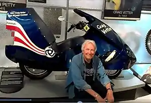 Craig Vetter, a gray haired man wearing a blue open shirt sitting just below his dark blue 1981 Streamliner motorcycle exhibited at a show in 2016