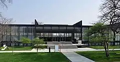 a low glass and steel building behind a sidewalk and small lawn and three trees