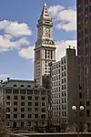 Distant, ground-level view of white limestone tower. The building is topped with a pyramidal-cap, and a large, 4-faced clock is visible just below the main roofline