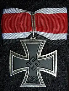 A silver framed black cross that has arms which are narrow at the center, and broader at the perimeter. In the middle of the cross is a swastika, an equilateral cross with its arms bent at right angles. On the lower arm of the cross are the number 1939 engraved. The cross is connected to a ribbon with a silver clip. The ribbon has a red central stripe, flanked in white and with a black edge stripe.