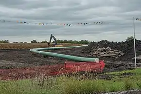 A long green pipeline is installed on a construction site with an excavator in the background