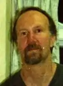 A middle-aged man with light brown hair and a goatee stands in front of a window