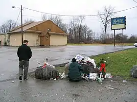 A standing man and sitting woman by a rock covered in flowers and other tributes to Dimebag Darrell Abbott. The Alrosa Villa nightclub is visible in the background.