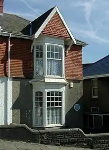 On a hill street stands a two-storeyed semi-detached house with bay windows to the front and a sloped tiled roof with a chimney.