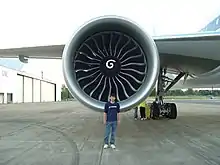  Aircraft engine, forward-facing view with a Boeing engineer in front to demonstrate the engine's size. The engine's large circular intake contains a central hub with a swirl mark, surrounded by multiple curved fan blades.