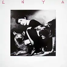 A black and white photograph of a brunette woman with a dark dress that is crouched with two wolves. The image is surrounded by a white border and the word "Enya", stylized in all capital letters, is placed on the top corner of the picture.