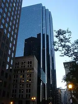 Ground-level view of a 40-story skyscraper with an all-glass facade and several setbacks. In the facade is a reflection of a dark brown building with lighter windows, and near the bottom of the skyscraper is a small white building with dark windows