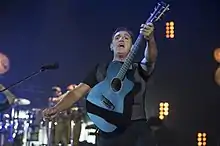 An image of a man wearing a black shirt and pants is performing while holding a black guitar