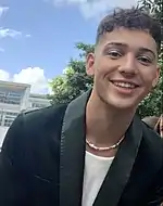 Man wearing a forest green suit and matching eyeshadow smiling towards the camera.