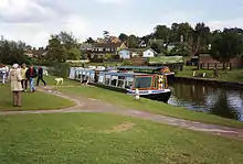 A narrow boat moored alongside grassy area. Houses can be seen in the background.