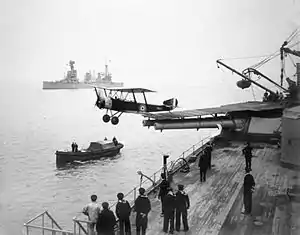 A small biplane has just taken off from a warship, having used a platform built over the roof and barrels of a twin-barrelled turret. Sailors are observing the launch from the deck of the ship, and from a small boat nearby. A large warship is visible in the background.