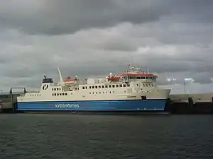 A large modern ferry with a blue hull and white topsides lies next to a harbour under grey skies.