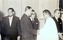 A smiling Babana, in white, greets men in suits.