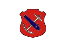 An insignia in the form of a red shield. On the shield are a white anchor crossed by a blue cannon barrel.