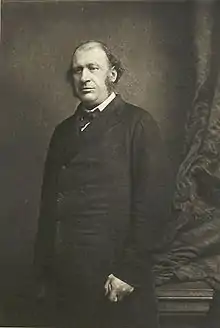 A black-and-white photograph of James Fitzjames Stephen, a balding man with sideburns. He stands looking to the left of the camera near a curtain.  He is holding a cane in his left hand.