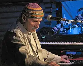 A man behind a microphone, with one of his hands on a black and white keyboard, wearing a dress shirt and a multi-colored cap.