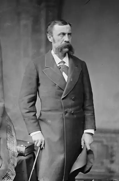 A man in his forties with black hair and a bushy, black mustache. He is standing, facing right, and holding a can in his right hand, a hat in his left, and wearing a long black coat