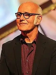 Ludovico Einaudi male wearing glasses and a black jacket, smiling and looking right of camera