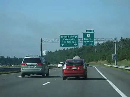 Ground-level view of three lanes of a divided expressway; two large green exit signs are visible in the distance, and the road is surrounded by dense forests.