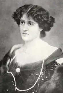 A white woman wearing a dark gown with a low neckline and a string of pearls; her dark wavy hair is parted center and dressed back to the nape