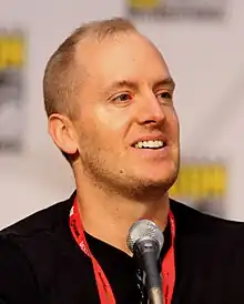 A man with a thin layer of brown hair sits behind a microphone.