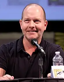 A man with closey shaven brown hair, speaking into a microphone, with his fist slightly raised above the table.