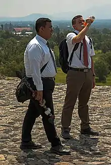 Two young men, the one on the left darker-skinned than the one on the right, who is drinking some orange liquid from a plastic bottle, standing on a stone surface with mountains in the background. They are both carrying backpacks and wearing white dress shirts, ties and slacks. The one on the right is wearing a short-sleeved shirt and tan slacks