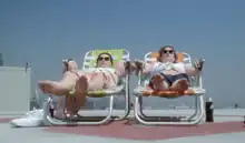 Two women with dark sunglasses sunbathe on the roof of a hospital.