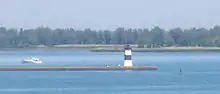 A pier with a square, black and white-striped lighthouse at the end. A small motorboat is paralleling the pier, heading away from the lighthouse.