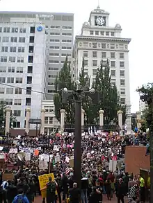 A tall, thin structure. In the background are tall buildings.  At its base, it is surrounded by crowds of people.