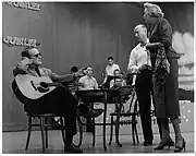 Red Foley sitting onstage at a table, left, with guitar on his lap; director Bryan Bisney and actress Fran Allison stand to the right