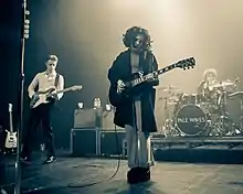 A woman stands in the foreground playing a guitar. A rhythm guitarist and drummer are in the background