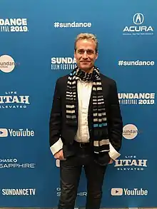 white male wearing black pants and jacket with white shirt and a scarf, standing with awards-ceremony stage background, smiling at camera