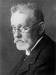  Portrait of an older, thin man with a beard wearing glasses and dressed in a suit and tie