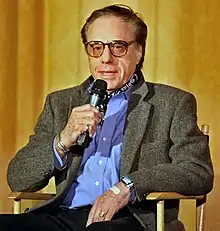 A white male in his 60s sitting in a chair while speaking into a microphone. He is wearing eyeglasses and a grey jacket over a blue buttoned down shirt. His left hand is rested on his lap.